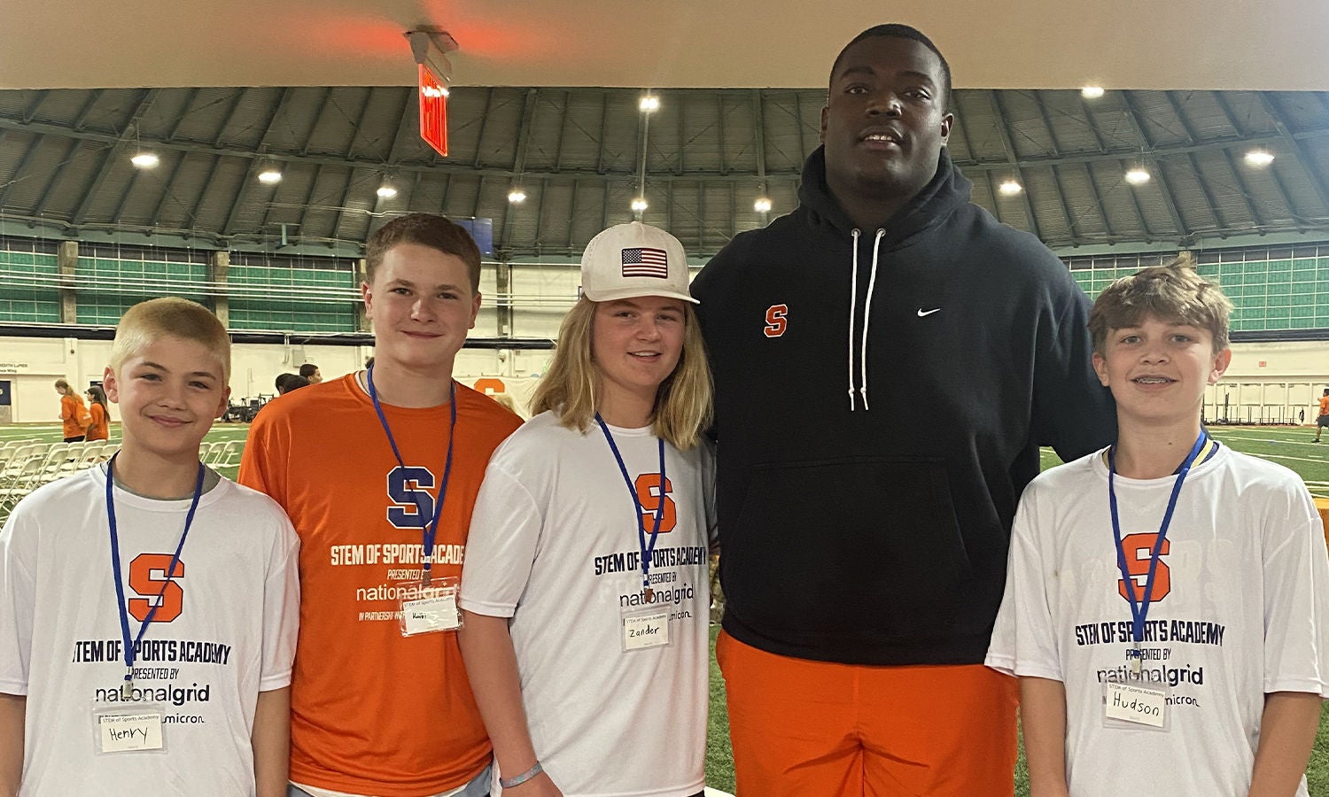 Five individuals participants pose on an indoor track field, four of whom are students wearing Stem STEM of Sports Academy' T-shirts